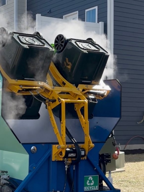 The trailer's yellow lift arm holding two wheeled bins upside down over the wash bay, steam rising around them