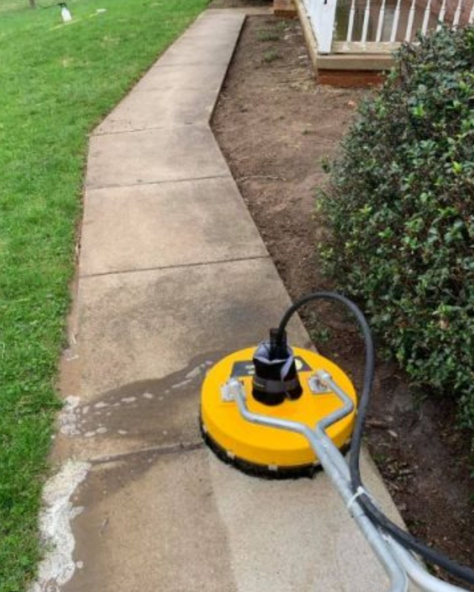 A yellow surface cleaner part-way along a concrete walkway, the strip it has already passed over visibly lighter than the concrete ahead of it