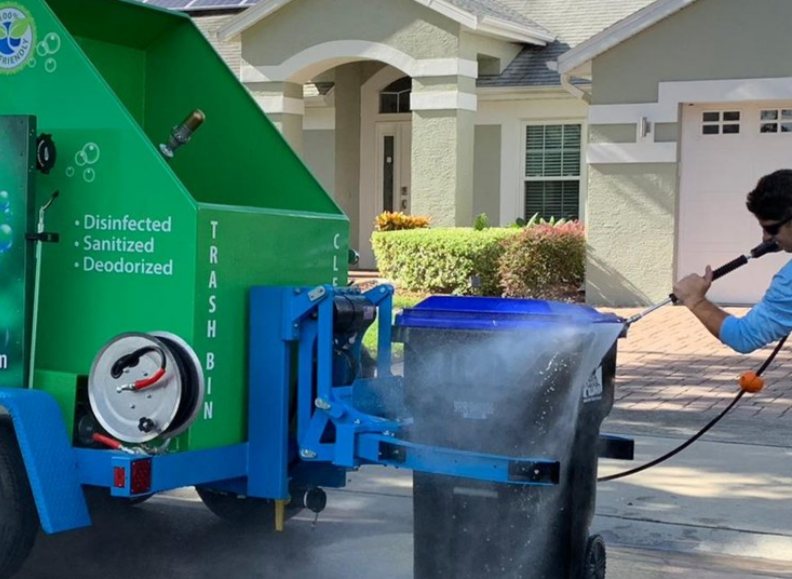 A NeatReason worker pressure-washing a wheeled trash bin clamped to the green and blue NeatReason trailer in a suburban driveway, spray and mist around the bin
