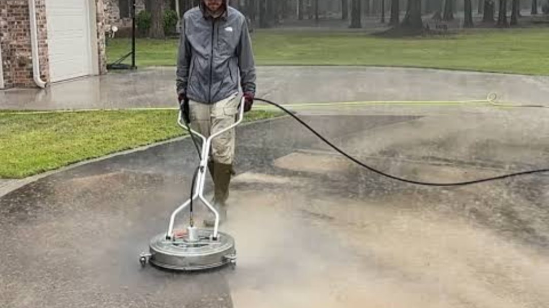 A NeatReason worker in a rain jacket walking a round surface cleaner across a wet driveway in the rain, a clean strip trailing behind him